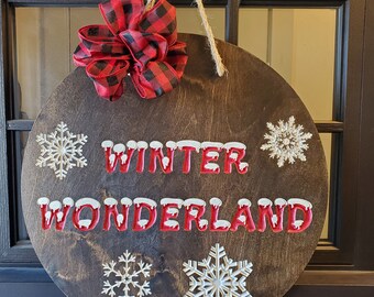 May include: A round wooden sign with the words "WINTER WONDERLAND" in red letters with white snow accents. The sign is decorated with snowflakes and a red and black buffalo plaid bow.