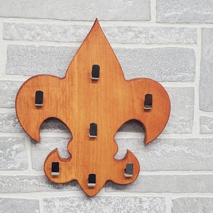 May include: A wooden wall-mounted fleur-de-lis shaped rack with seven metal hooks for hanging items.