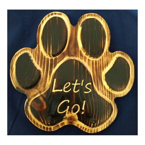 May include: A wooden paw print-shaped key holder with the words "Let's Go!" carved into the center. The wood has a dark, burnt finish with lighter, natural wood grain accents. Includes two small pegs for holding keys.