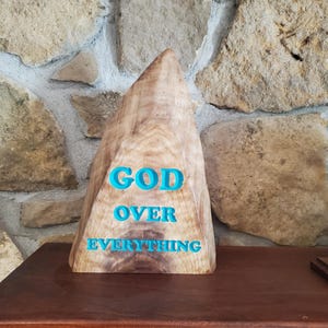May include: A wooden sculpture in the shape of a mountain with the words "GOD OVER EVERYTHING" carved into it in teal blue lettering.