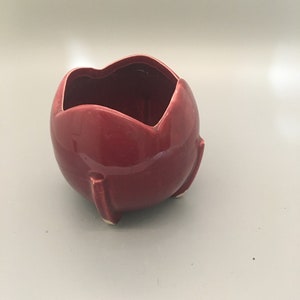 Puede incluir: Una maceta de cerámica de color burdeos brillante con una forma redondeada única. La maceta tiene un borde festoneado y tres pequeños pies redondeados. El interior es de un tono burdeos más oscuro.