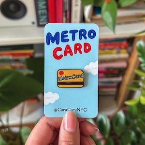 Può includere: Una spilla MetroCard azzurra con le parole "METRO CARD" in blu e rosso. La spilla presenta un design MetroCard giallo con le parole "MetroCard" e due disegni di nuvole bianche. Il testo "@CaraCaraNYC" è in basso.
