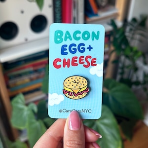 May include: A blue and white card with the text "Bacon Egg + Cheese" in colorful letters. A cartoon illustration of a bagel with bacon, egg, and cheese is on the card. The text "@CaraCaraNYC" is at the bottom of the card.