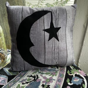 May include: A gray and black throw pillow with a crescent moon and a star design. The pillow has a woodgrain pattern.