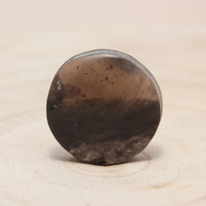 Peut inclure: Un bouchon de pierre rond, brun foncé, avec un effet de dégradé. La partie supérieure est d'un brun clair et translucide, passant à un brun plus foncé et opaque en bas. Les bords ont un léger éclat métallique. Le bouchon est posé sur un fond clair en bois.