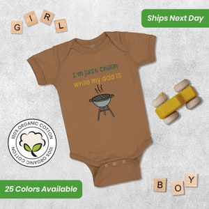 May include: A brown baby onesie with short sleeves and a white graphic that says "I'm just chillin' while my dad is" with a cartoon grill. The onesie is made of 100% organic cotton.