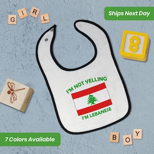 May include: White baby bib with a black border. The bib has a red, white, and green Lebanese flag with a cedar tree on it. The text "I'm not yelling I'm Lebanese" is printed on the bib.