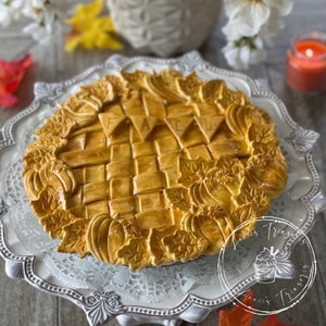 May include: A golden brown pie with a woven crust and fall-themed decorations. The pie is decorated with leaves, pumpkins, and the word "FALL" in banner flags. The pie is on a white cake stand with a lace doily.