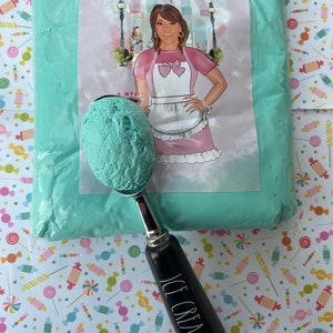 May include: A scoop of light blue ice cream in a silver ice cream scoop with a black handle that reads "ICE CREAM" in white. The scoop is in front of a bag of light blue material with a label that reads "Annie's Treasures". The background is a colorful candy pattern.