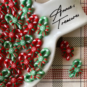 May include: A collection of red and green candy cane bow ornaments with white stripes. The ornaments are on a white ceramic serving dish with the text "Anne's Treasures" on the handle.