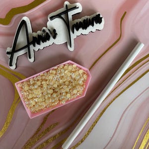 May include: A pink tray filled with light yellow rhinestones, a white pencil, and a sign that reads "Annie's Treasures" in black script. The background is pink with gold swirls.