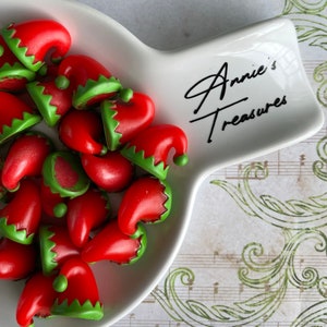 May include: A white, uniquely shaped dish filled with small, red elf hats with green trim. The dish has the text "Annie's Treasures" in black script. The background features a musical score and decorative green flourishes.
