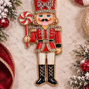 May include: A gold-tone metal nutcracker brooch featuring red and white enamel details, clear rhinestones on the hat, uniform, and belt, and a red and white peppermint candy held in one hand. The text Annie's Treasures is printed below.