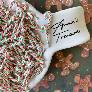 May include: A white ceramic dish filled with red and green candy canes. The dish has the words "Annie's Treasures" written on it. The dish is sitting on a green background with gingerbread men.