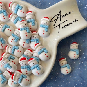 May include: A white ceramic plate filled with small, white snowman figurines with red hats and blue scarves. The plate is on a blue background with white dots. The text "Annie's Treasures" is written in black on the plate.