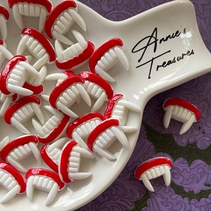 May include: White and red vampire teeth decorations arranged on a white, uniquely shaped plate. The teeth have a red gum line and are scattered across the plate. The plate has the text "Annie's Treasures" in black script.