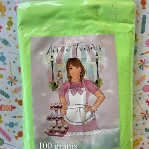 May include: A sealed plastic bag filled with bright neon green slime. The bag has a label with the text "Annie's Treasures" and an illustration of a woman in a pink apron. The label also states "100 grams".