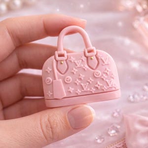 May include: A small, pink handbag-shaped object with a handle, embossed with a repeating pattern of stars and the letters "LV". The bag has gold-colored accents and a small tag. The background is a soft pink with pearls and other decorative elements.