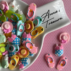 May include: A collection of miniature summer-themed embellishments, including pink, yellow, and blue flip-flops, sun hats, and swimsuits. The items are arranged on a white dish with the text "Annie's Treasures" in black script, set against a pink polka-dot background.