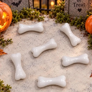 May include: A collection of six smooth, white, bone-shaped objects arranged on a speckled surface. The scene includes two miniature tombstones, one marked with RIP and a bat, the other with Trick or Treat, alongside a glowing orange pumpkin.
