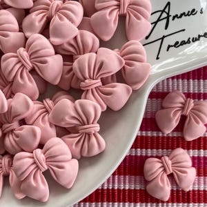 May include: A collection of light pink bow-shaped embellishments. Some bows are on a white, curved plate with the text "Annie's Treasures". The bows are scattered on a red and white checkered surface.