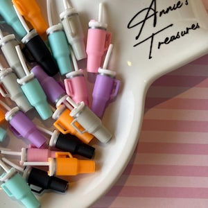 May include: A collection of colorful miniature tumblers with lids and straws. The tumblers are in various colors, including pink, purple, blue, green, and gray. The tumblers are arranged on a white plate with a pink and white striped background. The plate has the text "Annie's Treasures" written on it.