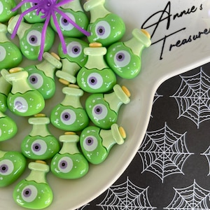 May include: A collection of green potion bottle-shaped charms with eyeball designs, arranged on a white plate. A purple spider is placed on top of the charms. The plate is on a black surface with a white spiderweb pattern. The plate has the text "Annie's Treasures".