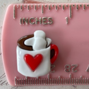 May include: A white ceramic mug with a red heart on the front, filled with brown liquid and topped with a white marshmallow. The mug has a brown rim and a red handle.