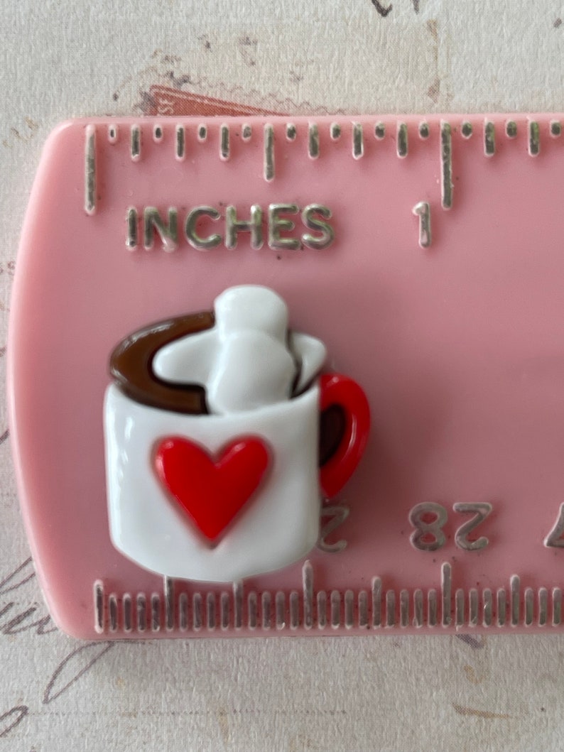May include: A white ceramic mug with a red heart on the front, filled with brown liquid and topped with a white marshmallow. The mug has a brown rim and a red handle.