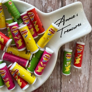May include: Miniature Lay's Stax potato chip containers in various colors, including red, yellow, green, and pink, are arranged in a white, uniquely shaped dish. The dish is inscribed with "Annie's Treasures" in cursive. The chips are a snack food.