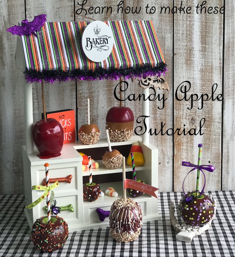 K&ouml;nnte beinhalten: Ein wei&szlig;er Holzstand f&uuml;r Apfel-Karamellen mit einem gestreiften Vordach und einem Schild mit der Aufschrift "Bakery". Der Stand ist mit Halloween-Dekorationen wie Flederm&auml;usen und Maisbonbons geschm&uuml;ckt. Es werden mehrere Apfel-Karamellen ausgestellt, die jeweils mit verschiedenen Toppings wie Streuseln, Schokolade und Karamell verziert sind. Der Text "Candy Apple Tutorial" ist auf dem Stand geschrieben.