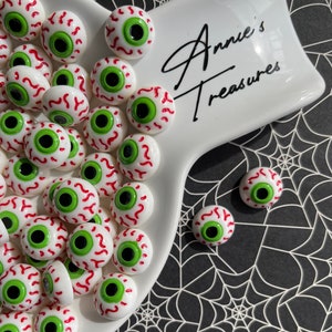 May include: A white ceramic dish with a black inscription "Annie's Treasures" filled with numerous small, white, red, and green googly eyes.  A few googly eyes are scattered on a black and white spiderweb background.