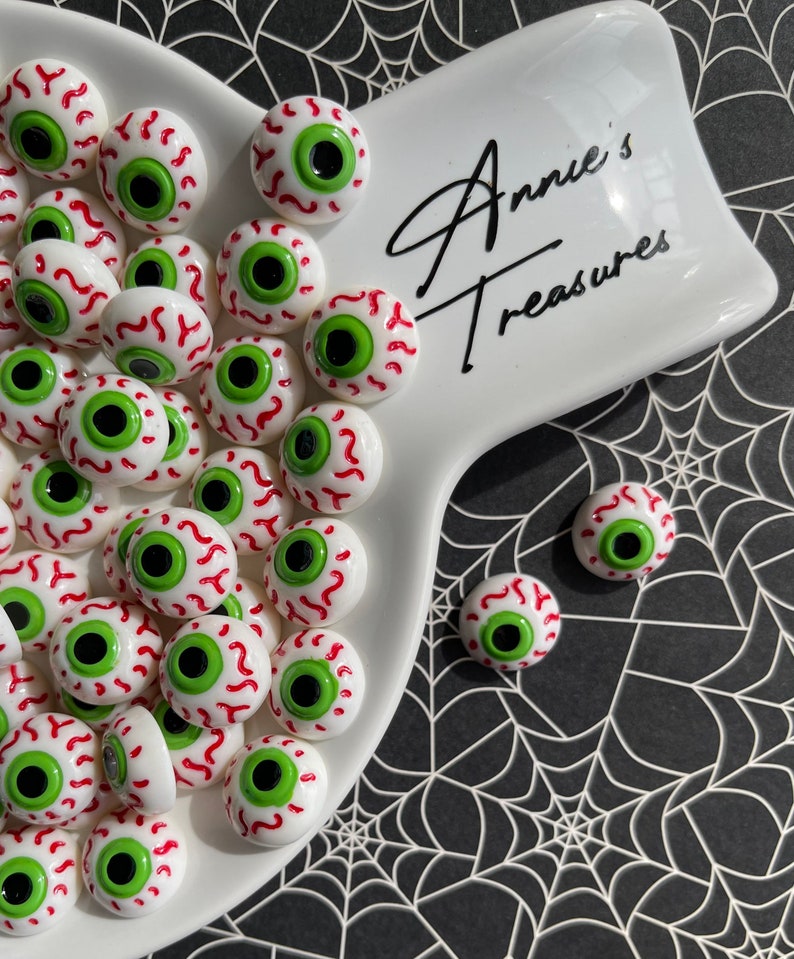 May include: A white ceramic dish with a black inscription "Annie's Treasures" filled with numerous small, white, red, and green googly eyes.  A few googly eyes are scattered on a black and white spiderweb background.