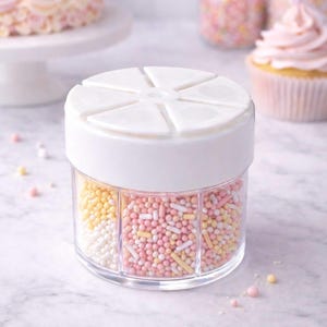 May include: A clear, segmented container filled with colorful sprinkles. The sprinkles are divided into sections of white, yellow, and pink. A white lid sits on top. A cake and cupcake are in the background.