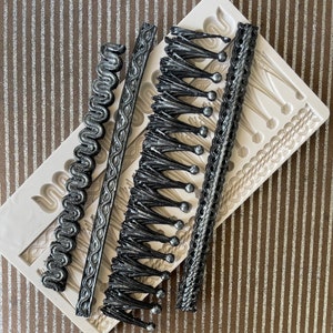 May include: A silicone mold for making decorative cake borders. The mold features five different designs, including a tassel, a rope, a swirl, and two different types of trim. The mold is white and the designs are black.