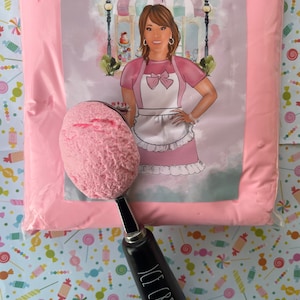 May include: A pink bag of frosting with a scoop of pink frosting on a black ice cream scoop. The bag has a label with a cartoon illustration of a woman in a pink dress and apron, with the text "Annie's Treasures". The background is a blue and white pattern of candy.
