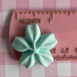 May include: A light blue plastic flower with five petals, measuring approximately 1 inch in diameter, is placed on a pink ruler with the words "INCHES" and "1" visible.