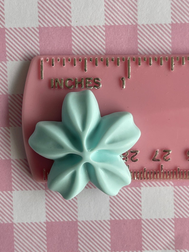 May include: A light blue plastic flower with five petals, measuring approximately 1 inch in diameter, is placed on a pink ruler with the words "INCHES" and "1" visible.