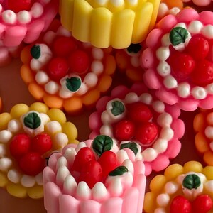 以下が含まれることがあります： 白いフロスティングと赤いイチゴをトッピングしたミニケーキの詰め合わせ。ケーキは緑の葉で飾られており、黄色、ピンク、またはオレンジのベースがあります。