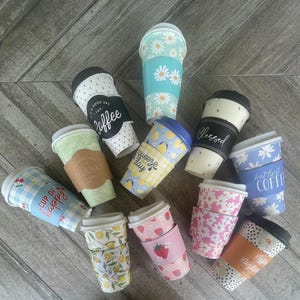 May include: An assortment of disposable coffee cups with lids and decorative sleeves. Designs include floral patterns, strawberries, lemons, and text like "Blessed" and "Cup of Happy". The cups are in various colors, including blue, pink, and green.