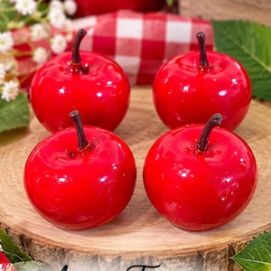 May include: Four glossy, bright red artificial apples with dark brown stems arranged in a square formation. The text "Annie's Treasures" is printed in a black cursive font at the bottom of the image, centered below the round wooden base.