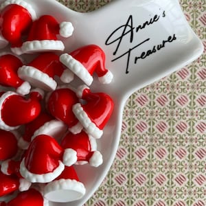 May include: A white ceramic dish with a red and white Santa hat design. The dish has the text "Annie's Treasures" written on it. The dish is filled with small red and white Santa hats.