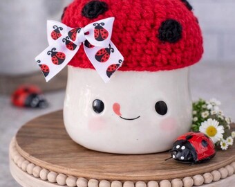 May include: A whimsical ceramic mug with a ladybug design. The mug features a red crocheted ladybug hat with black spots and a white bow with ladybug prints. The mug is on a wooden stand, with ladybug decorations.