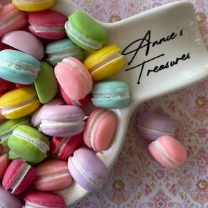 May include: A collection of pastel-colored macarons in a white ceramic dish with the text "Annie's Treasures". The macarons are arranged in a variety of colors, including pink, blue, yellow, and green.