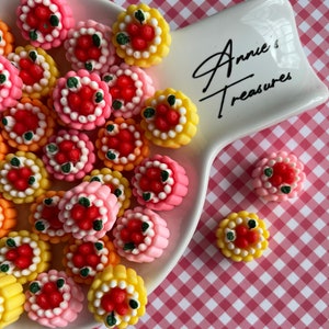 以下が含まれることがあります： 取っ手付きの白い陶器の皿に、小さくカラフルな装飾的なケーキが盛り付けられています。ケーキは赤と緑のアクセントで飾られており、イチゴと葉に似ています。皿には「Annie's Treasures」という文字が書かれています。