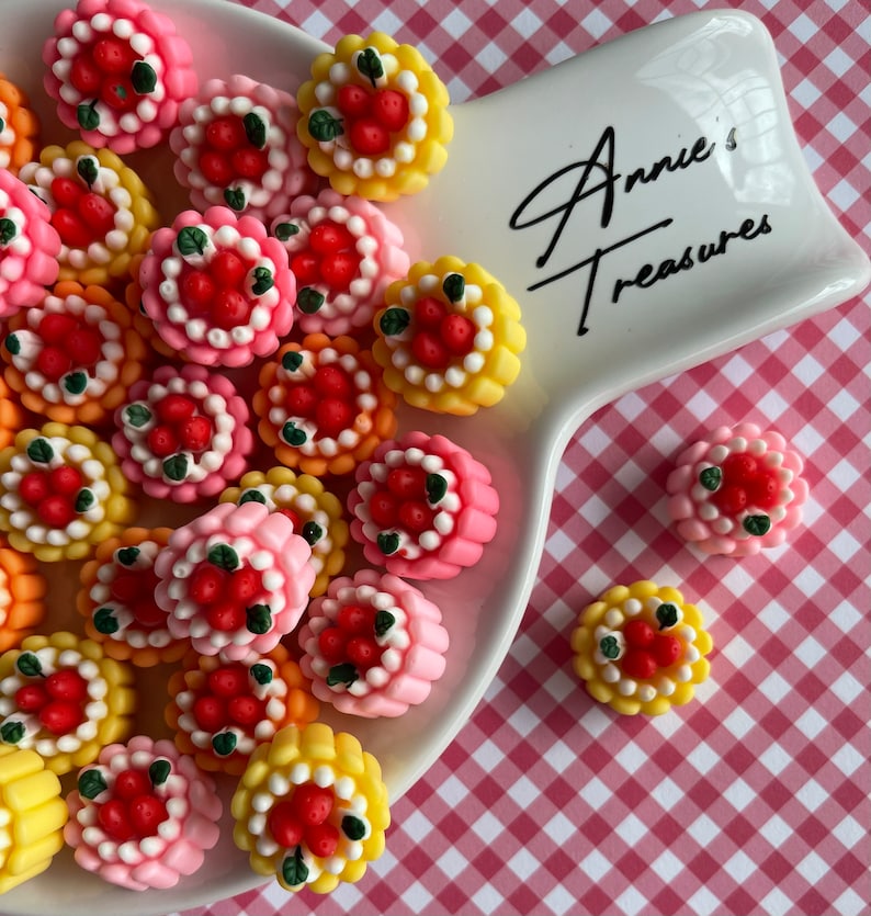 以下が含まれることがあります： 取っ手付きの白い陶器の皿に、小さくカラフルな装飾的なケーキが盛り付けられています。ケーキは赤と緑のアクセントで飾られており、イチゴと葉に似ています。皿には「Annie's Treasures」という文字が書かれています。