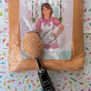 May include: A bag of brown ice cream mix with a scoop of ice cream in a silver scoop with a black handle that says "ICE CREAM". The bag is in front of a white card with a cartoon of a woman with a pink bow and the text "Annie's Treasures".
