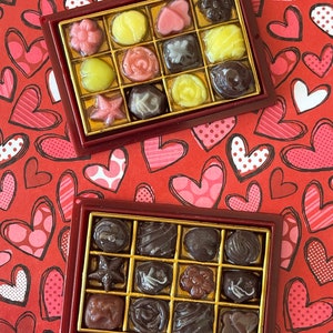 May include: Two boxes of chocolate candies. The top box is red with gold trim and contains a variety of chocolates with different fillings. The bottom box is red with gold trim and contains a variety of dark chocolate candies.