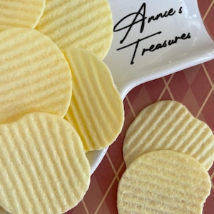 May include: Close-up of a pile of ridged, pale yellow potato chips. The chips are arranged on a white dish with the text "Annie's Treasures" in black script. The background is a red and white diamond pattern.