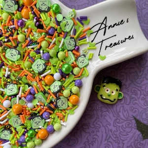 May include: A white ceramic dish filled with green, orange, and purple Halloween sprinkles. The sprinkles are shaped like Frankenstein's face and include small balls and sticks. The dish is on a purple background with the text "Annie's Treasures" on the side.